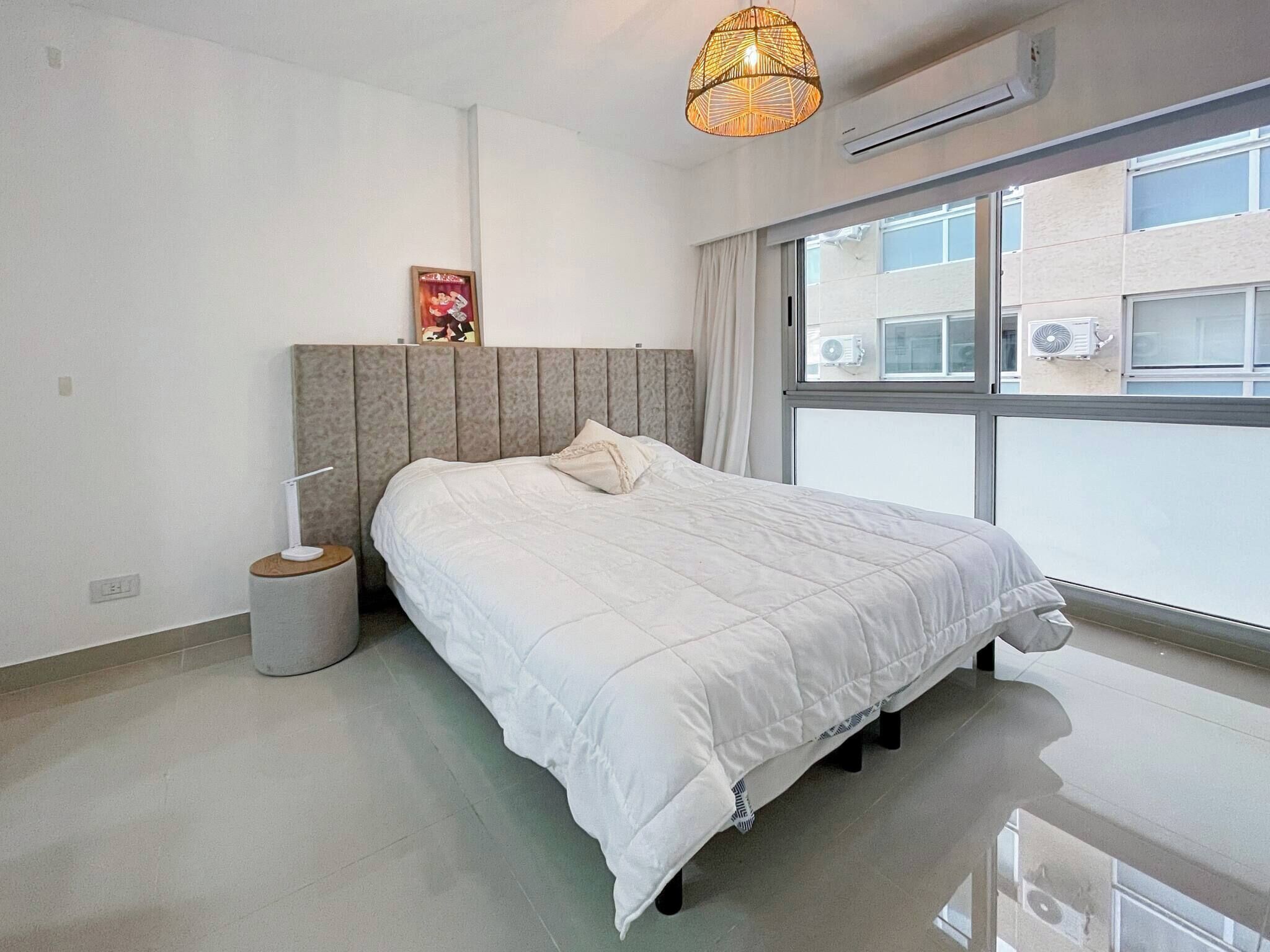 A modern bedroom with a large bed, a bedside lamp, a waste bin, and a window with an air conditioning unit.