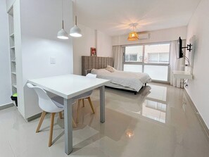 Ein modernes Schlafzimmer mit Bett, Schreibtisch und Stuhl. Ein Fenster mit Vorhängen, ein Deckenventilator und ein an der Wand befestigtes Fernsehgerät sind ebenfalls vorhanden.