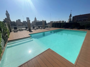 Ein Pool auf dem Dach mit Holzterrasse und Liegestühlen, umgeben von Stadtgebäuden und einem klaren Himmel.
