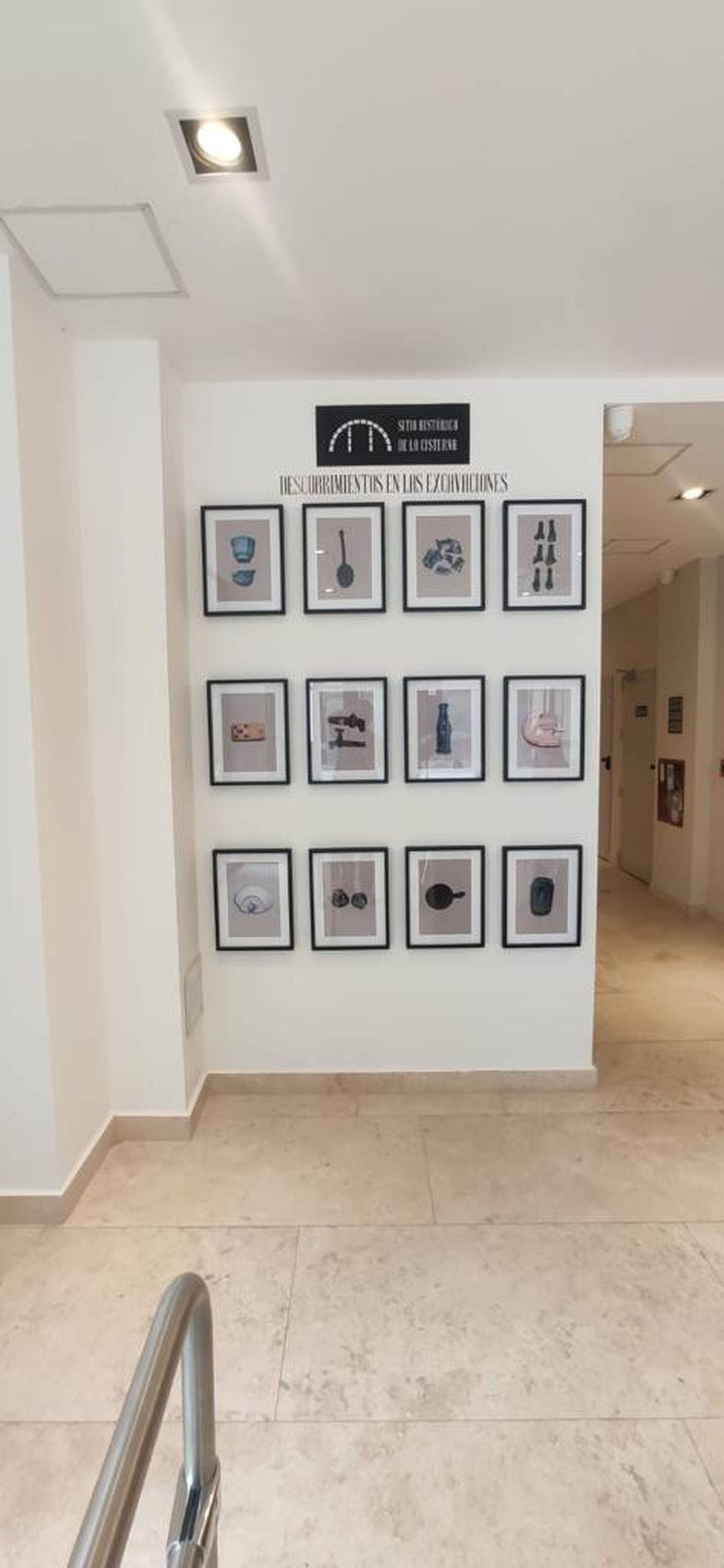 Un pasillo con una serie de obras de arte enmarcadas en la pared, un cartel encima y un suelo de baldosas.