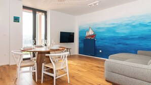 A modern living room with a wooden floor, a grey sofa, a dining area with a round table and white chairs, and a large wall mural of the ocean. - Bellavista Apartment Koper (Koper)