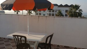 Une terrasse avec une table et des chaises, un parasol et une vue sur un bâtiment bordé de palmiers.