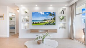 Un salon moderne avec un téléviseur à écran plat fixé au mur, une table basse en marbre et une vue sur la plage, visible à travers une grande fenêtre.