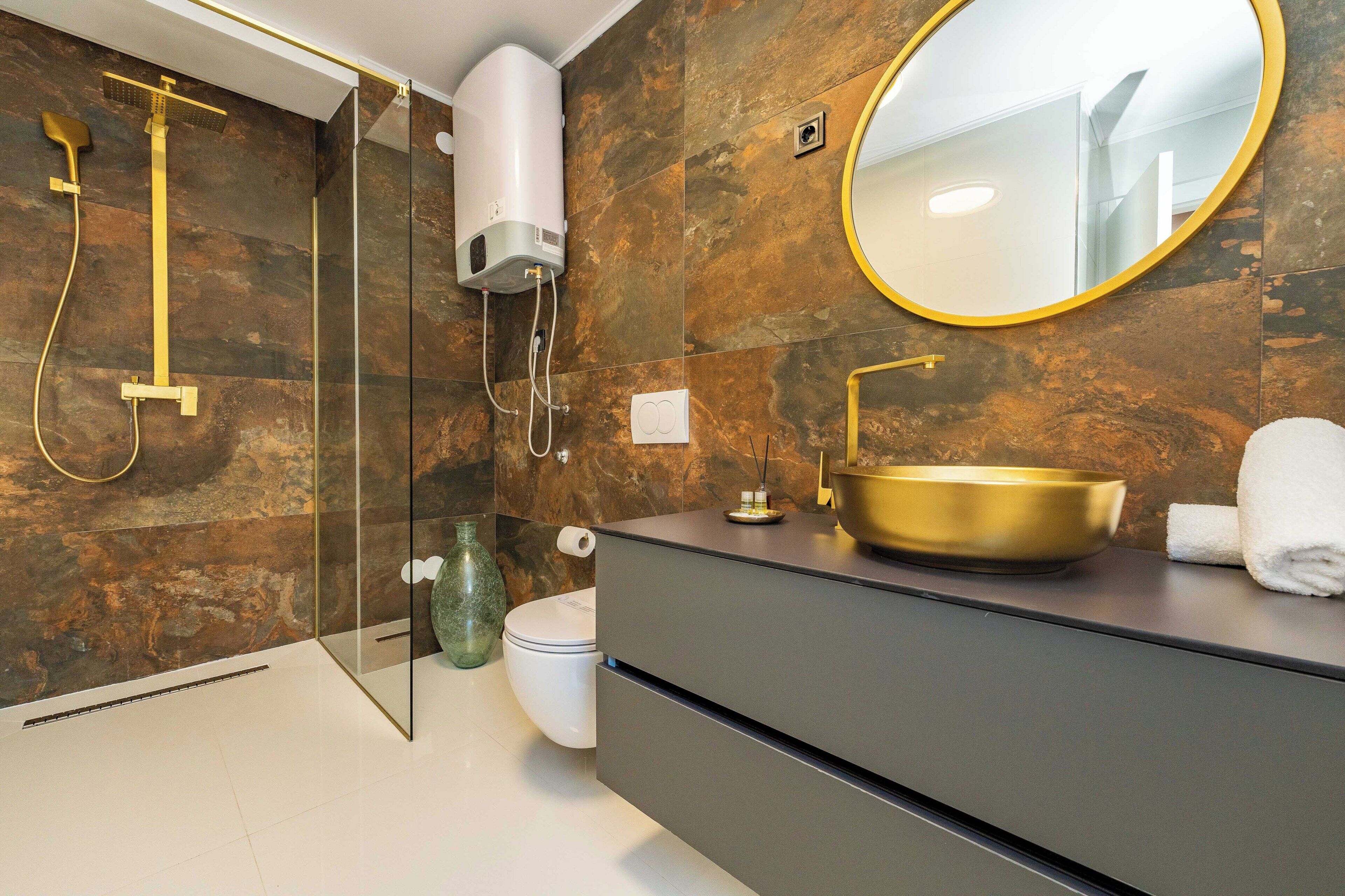 A modern bathroom with a glass-enclosed shower, a gold sink, and a round mirror.