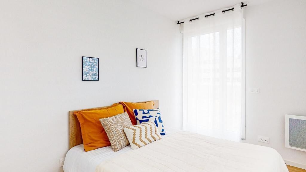 A neatly made bed with a white comforter and several pillows, a white wall with two framed pictures, and a window with white curtains.