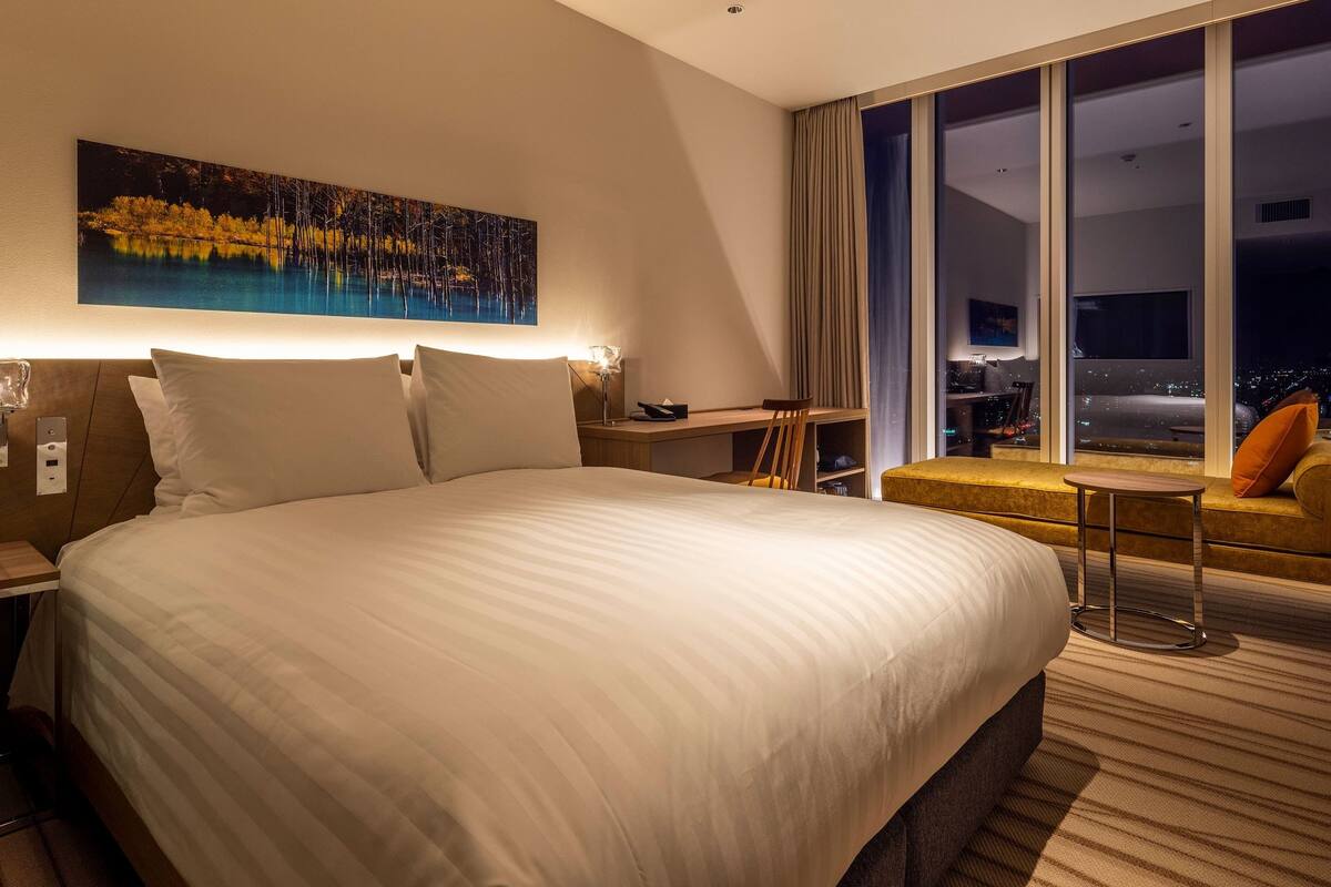 A hotel room with a large bed, a desk, a chair, and a view of the city at night.