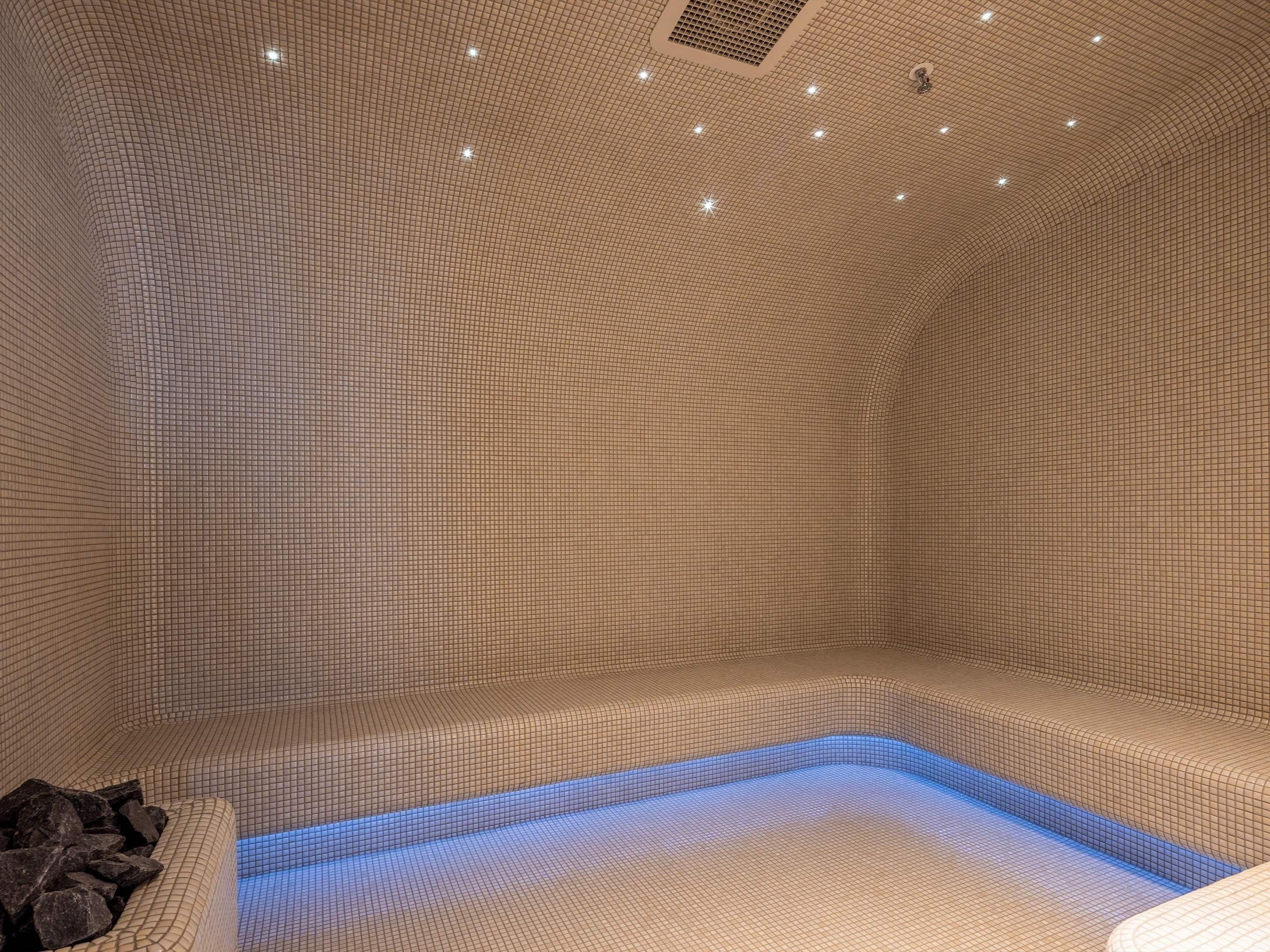 A spa room with a tiled floor and walls, a blue-lit jacuzzi, and a rock feature.