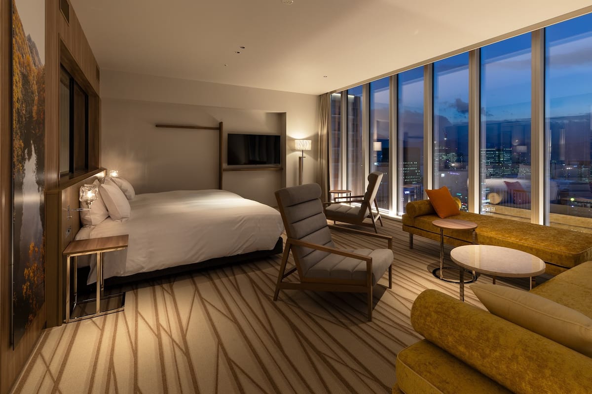A modern hotel room with a large bed, a seating area with a sofa and armchairs, a TV mounted on the wall, and a city view through large windows.
