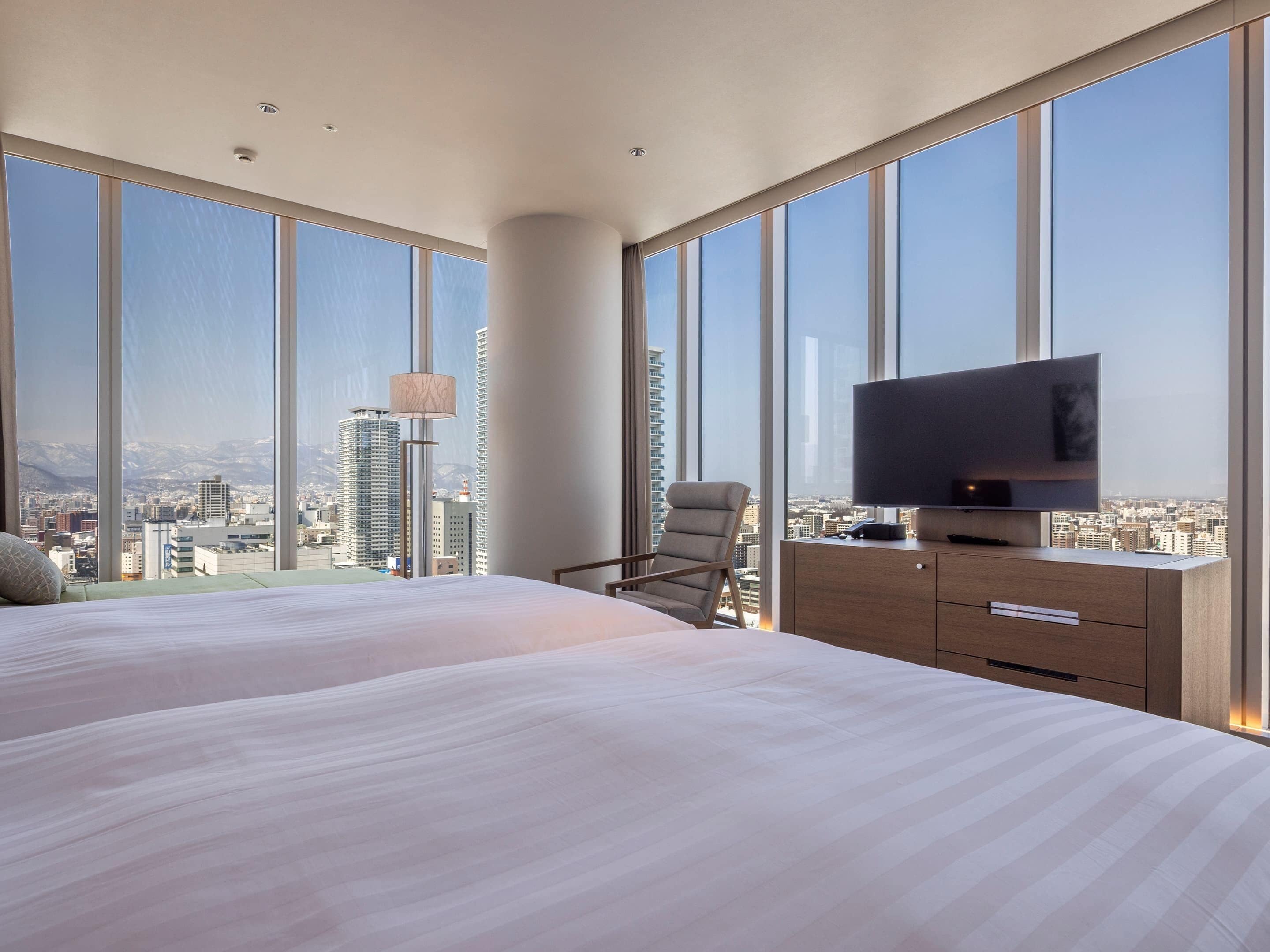 A modern hotel room with large windows offering a city view, a flat-screen TV, a bed, a chair, and a wooden dresser.