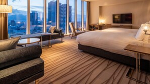 Habitación de hotel con una cama grande, un sofá, una mesita y un televisor. La habitación tiene ventanas amplias con vistas a la ciudad.