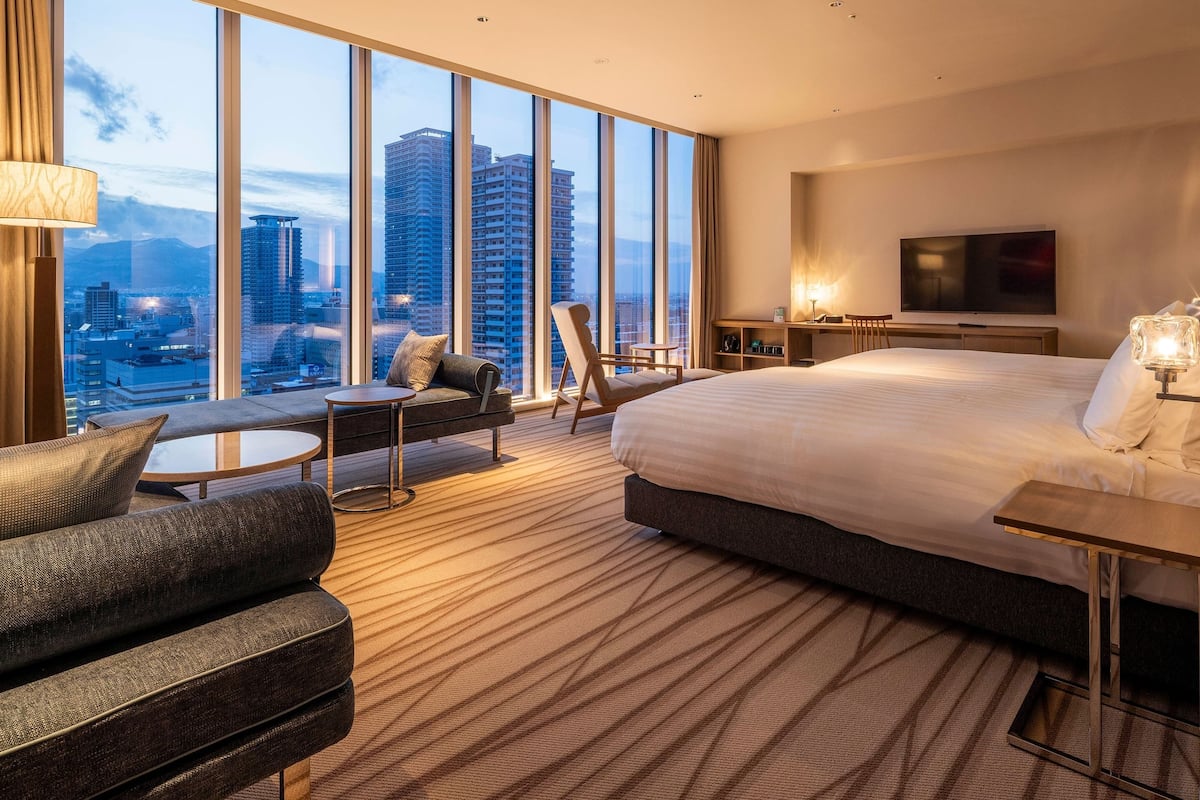 A hotel room with a large bed, a sofa, a small table, and a TV. The room has large windows with a city view.