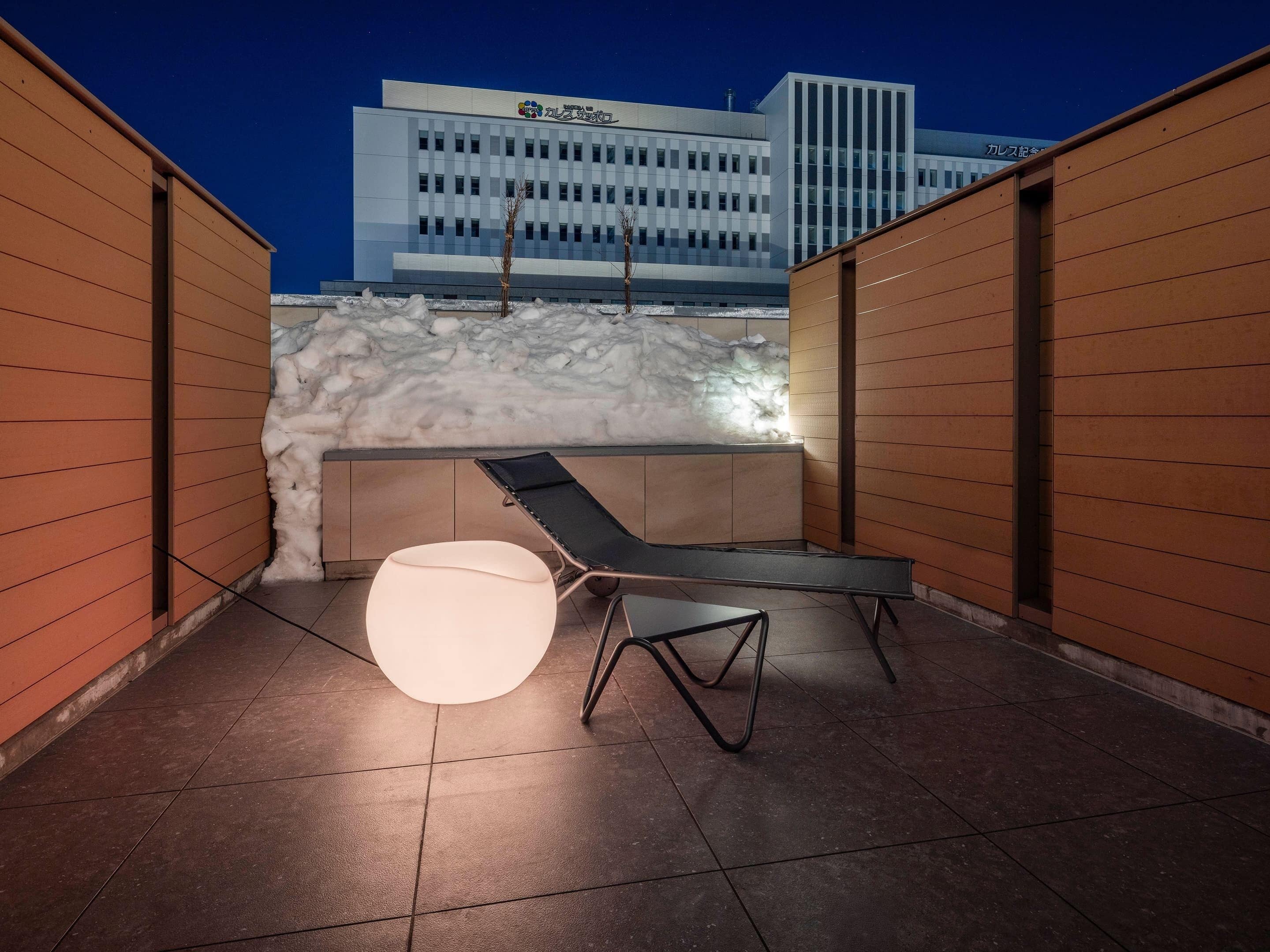 En modern utomhusplats med sittmöbler, inklusive en fåtölj och ett runt bord, en snötäckt vägg och en byggnad med skyltar i bakgrunden.