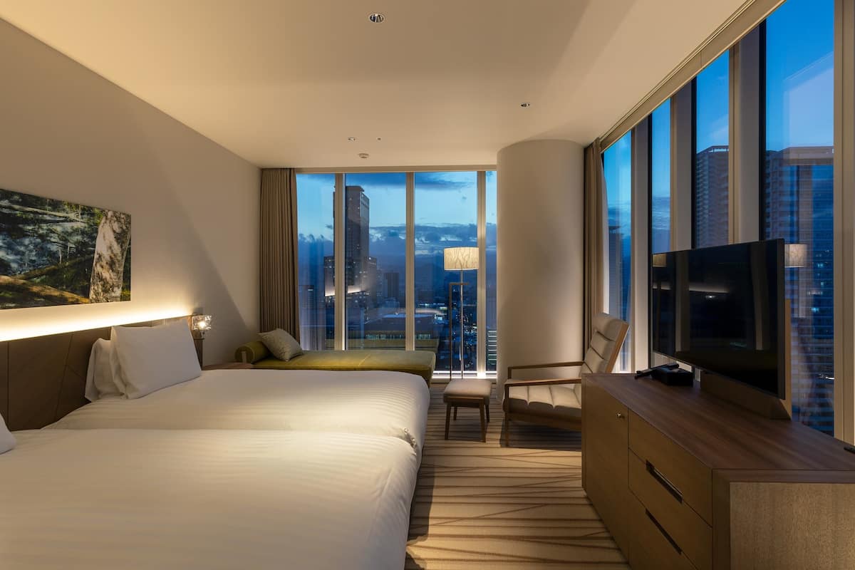 A modern hotel room with two beds, a TV, a desk, and a large window offering a city view.