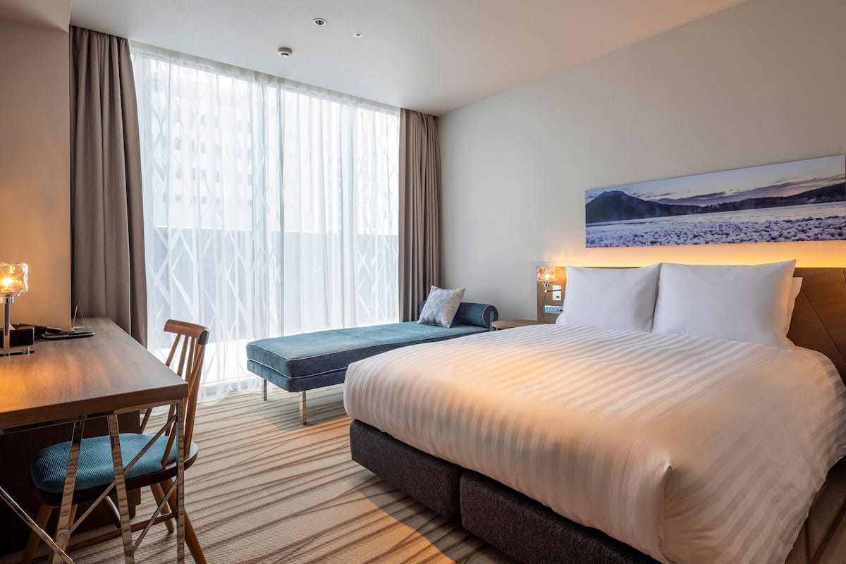 A hotel room with a large bed, a desk with a chair, a sofa, and a large window with curtains.