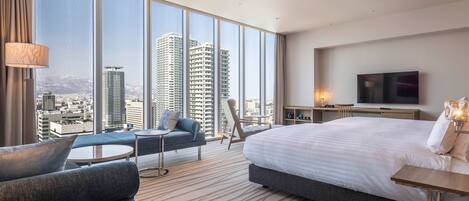 Una habitación de hotel moderna con una cama grande, un sofá, una mesita, un televisor y vistas a edificios altos.
