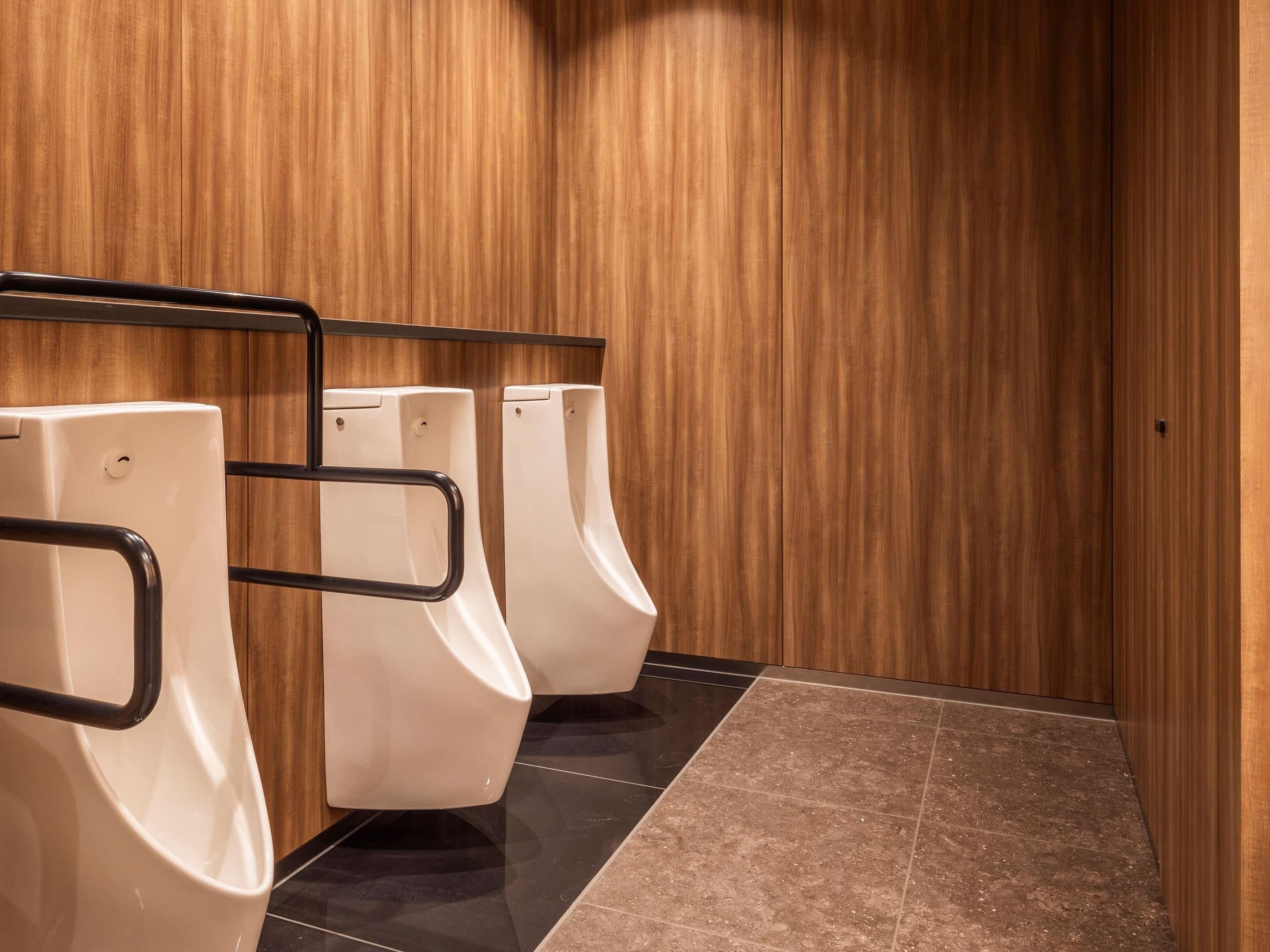A public restroom with three urinals against a wooden wall, a black tiled floor, and a metal handrail.