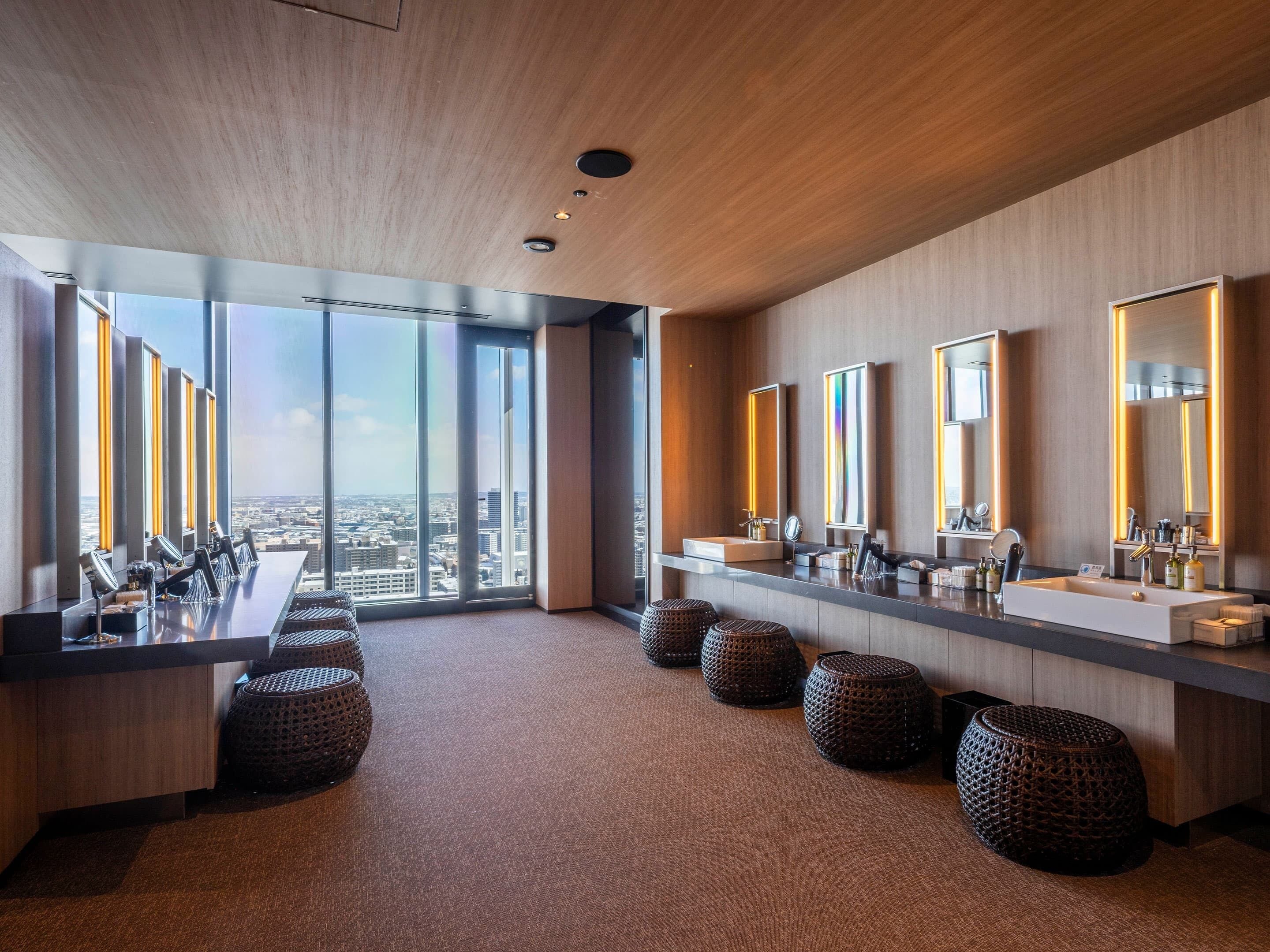 A modern hotel bathroom with large mirrors, a long vanity, and a city view.