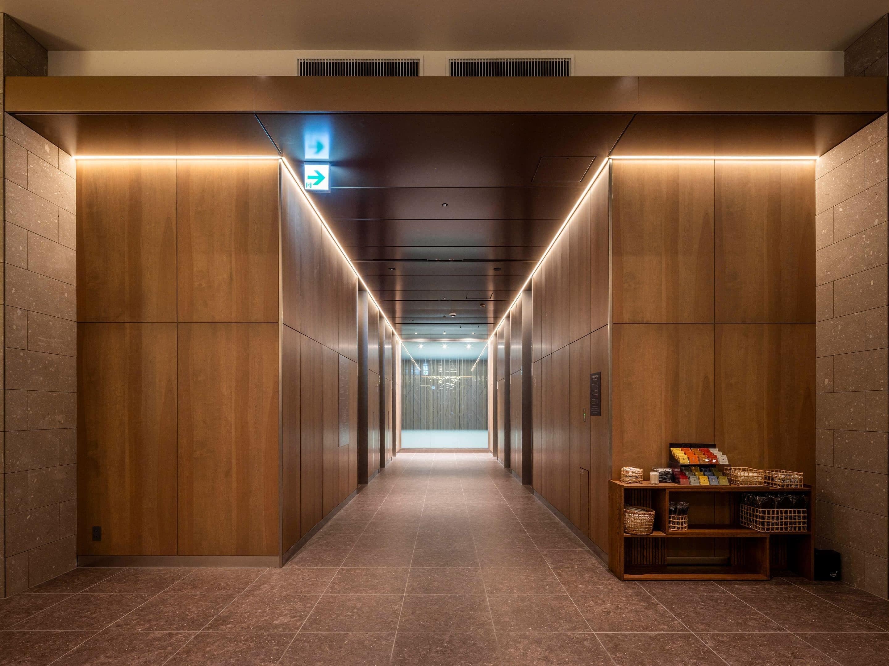 Un hall d’ascenseur moderne avec des murs en bois et un sol carrelé, qui mène à une sortie lumineuse.