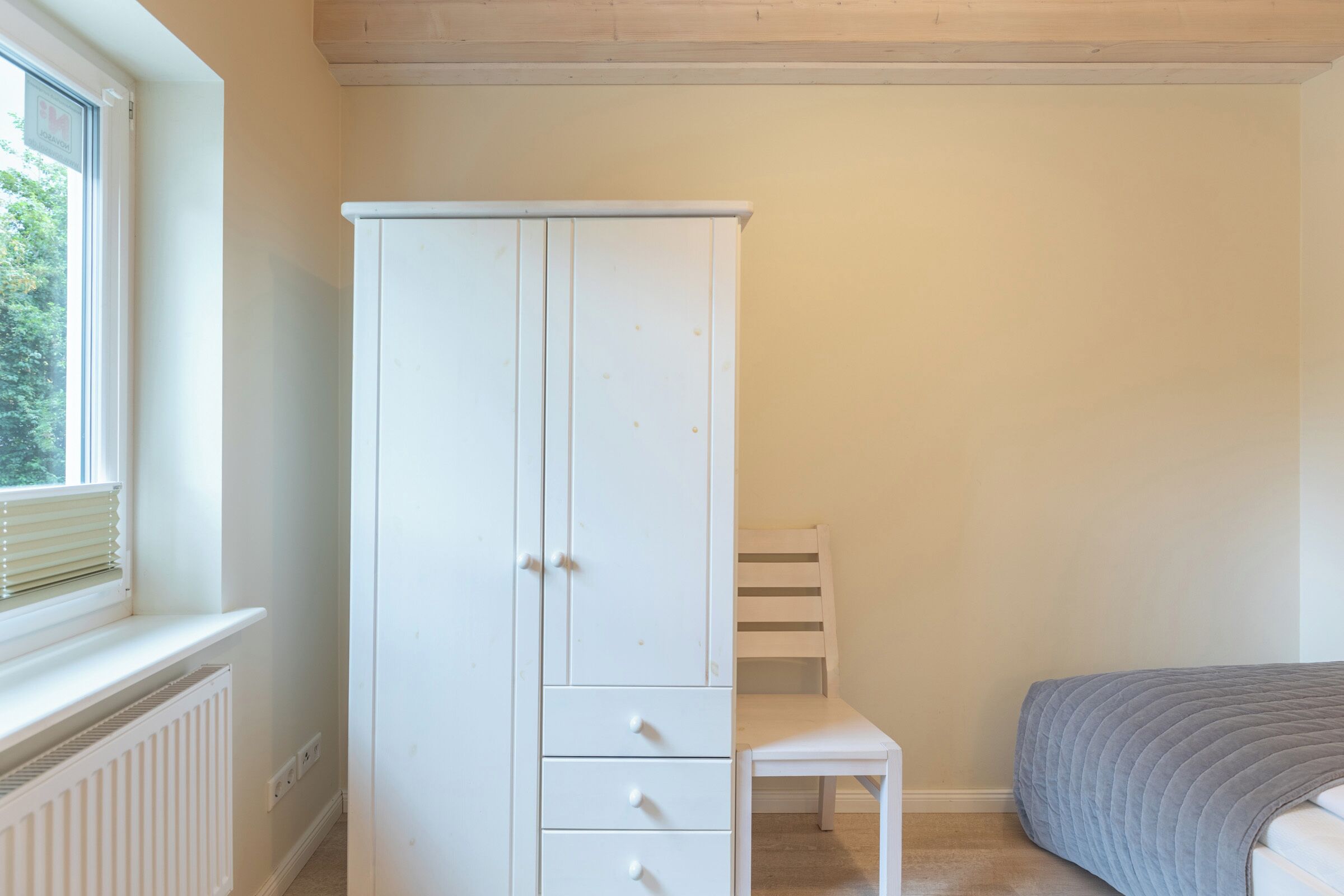 A room with a white wardrobe, a small white chair, and a bed with a grey quilt.
