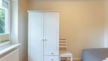 A room with a white wardrobe, a small white chair, and a bed with a grey quilt.