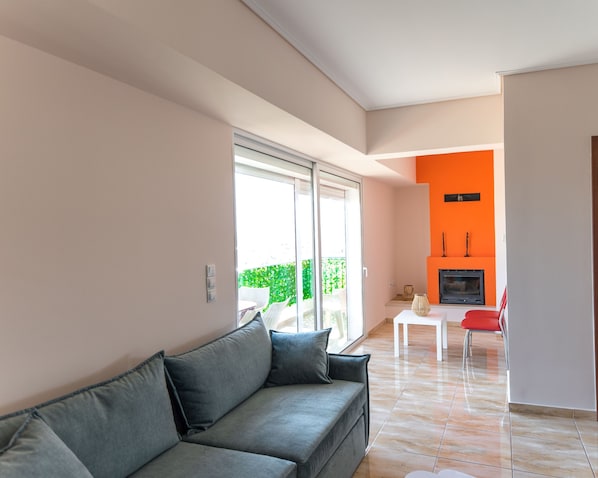 A modern living room with a grey sofa, a white coffee table, and a red chair. There is a glass door leading to a balcony with green plants.