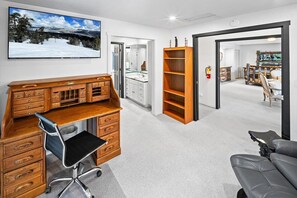 A room with a wooden desk, a black office chair, a bookshelf, a sofa, and a large window with a view of trees and mountains.