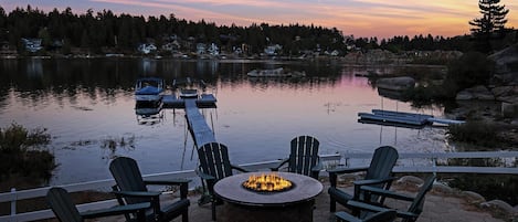 Terraza junto al lago con sillas Adirondack, un brasero y un muelle con barcos.