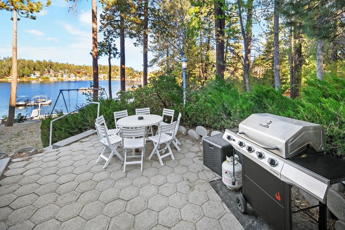 An outdoor patio with a grill, white chairs, and a view of a lake.