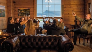 Un gruppo di persone brinda con bicchieri di vino in un ambiente accogliente e in legno, con una grande finestra che si affaccia su un paesaggio suggestivo.