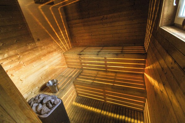 A sauna with wooden walls and benches, a metal bucket, and a stone-filled container. - Bjálki Cabin Lodge (Fjardabyggd)