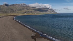 Una spiaggia con un litorale roccioso, un piccolo insediamento in lontananza e una montagna con le nuvole.