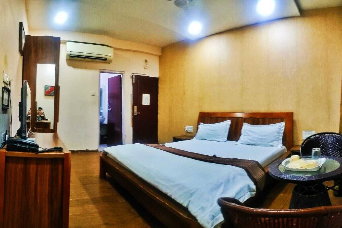 A hotel room with a double bed, a small table with a chair, a desk with a computer, and an air conditioning unit.
