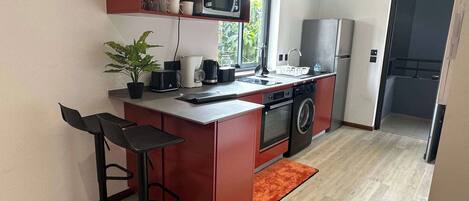 A compact kitchen with a microwave, toaster, coffee maker, and washing machine. There are two bar stools and a small potted plant.