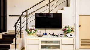 A modern living room with a flat-screen TV mounted on a white console, a staircase with black railings, and two flower arrangements on the console. - Value Inn Homestay by uBook (Butterworth)