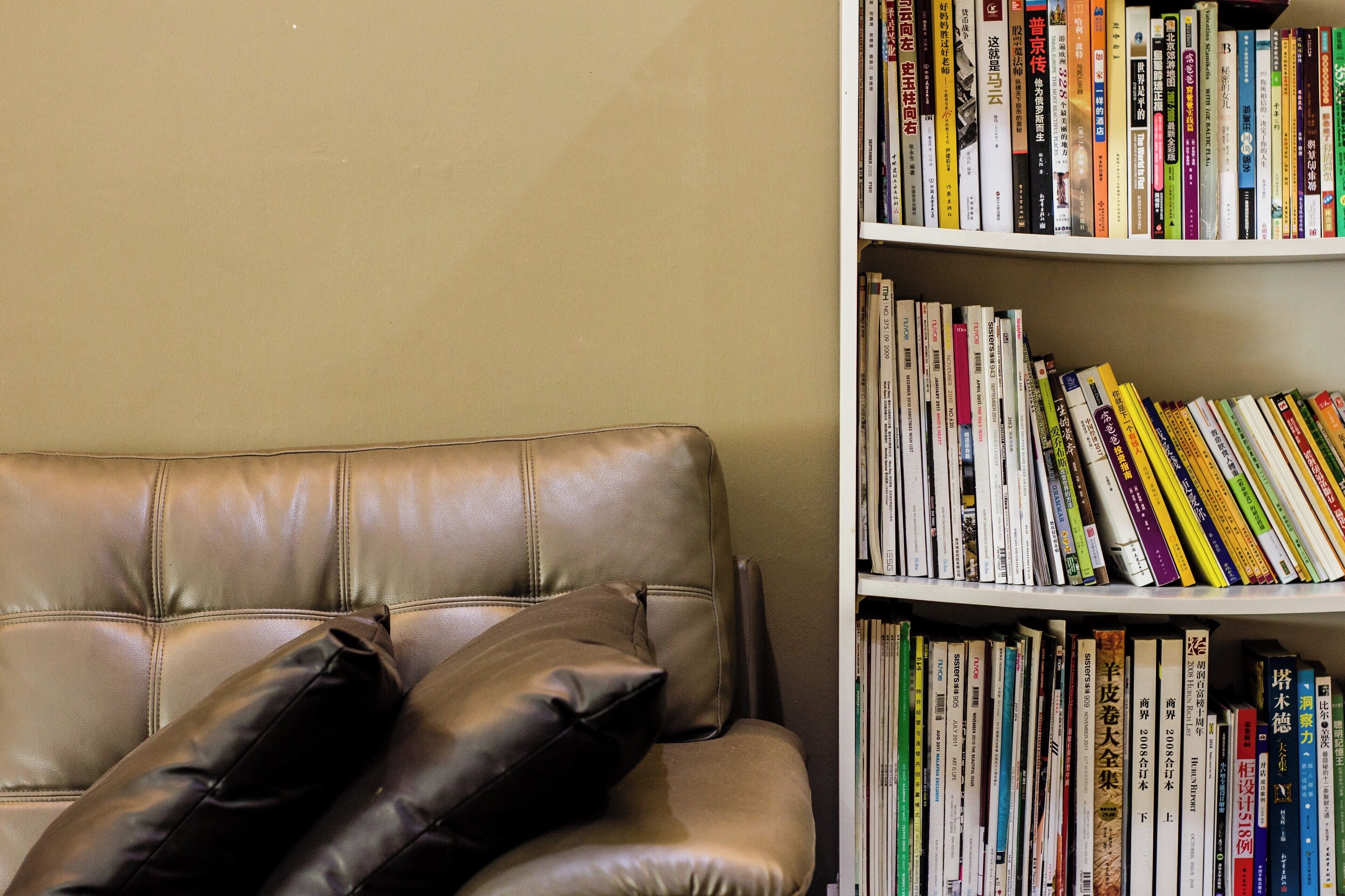 A leather sofa with black cushions against a beige wall, next to a bookshelf filled with books.