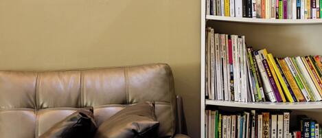 A leather sofa with black cushions against a beige wall, next to a bookshelf filled with books.
