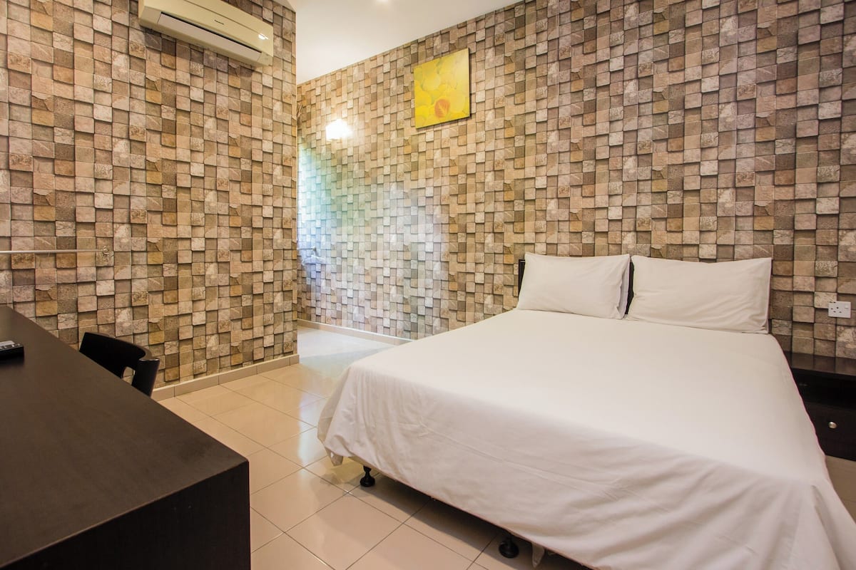 A hotel room with a stone-patterned wall, a bed with white bedding, a desk with a chair, and an air conditioning unit.