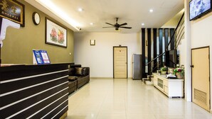 A hotel reception area with a reception desk, a sofa, a clock, a ceiling fan, a staircase, and a TV on the wall. - Value Inn Homestay by uBook (Butterworth)