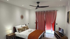 A bedroom with a bed, a nightstand, a TV stand, a ceiling fan, and a window with curtains. - Superbe T1 a 50 metre de la mer (Cayenne)