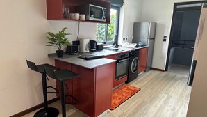 A compact kitchen with a microwave, oven, and washing machine. There are two bar stools and a potted plant. - Magnifique T1 avec piscine (Cayenne)