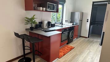 Eine kompakte Küche mit Mikrowelle, Toaster, Kaffeemaschine und Waschmaschine. Zwei Barhocker und ein kleiner Topfpflanze sind ebenfalls vorhanden.