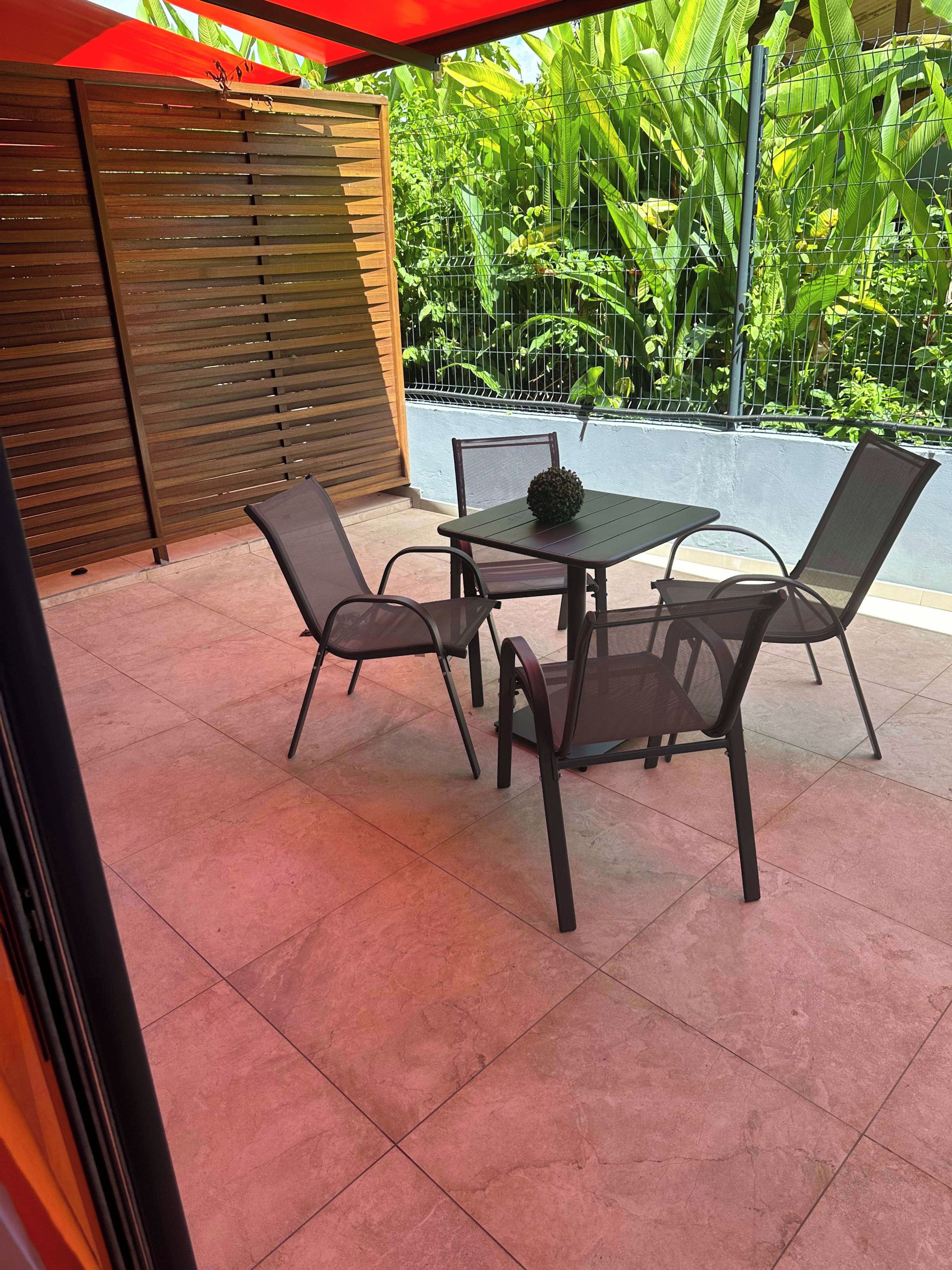 Um pátio externo com mesa e cadeiras, cercado por um muro de madeira e vegetação exuberante.