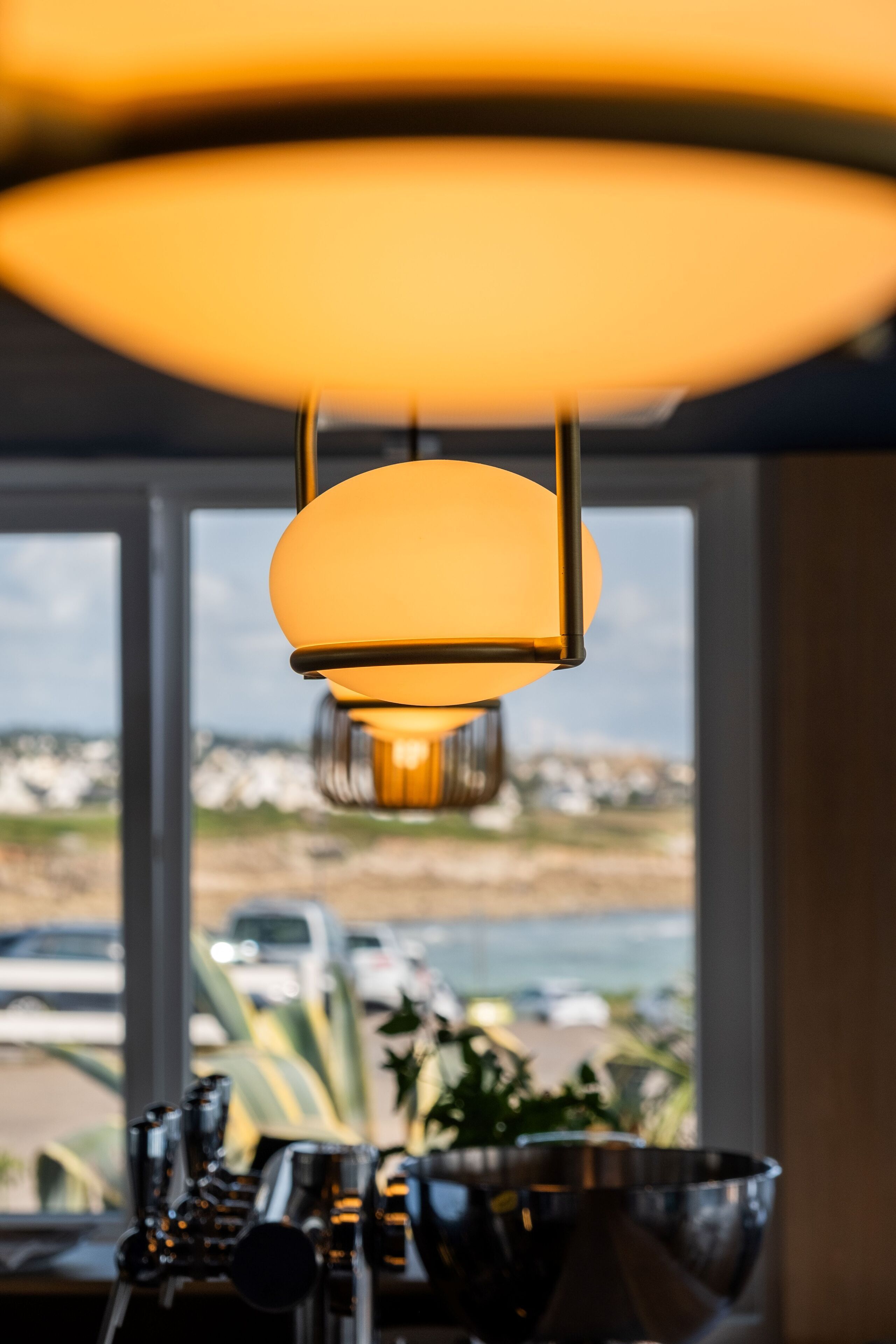 Un gros plan d’une lampe suspendue diffusant une lumière chaleureuse, une fenêtre donnant sur un parking et des ustensiles de cuisine posés sur un plan de travail.