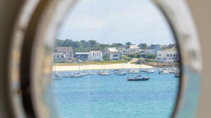Une vue à travers une fenêtre circulaire sur une zone côtière, avec des bateaux, des bâtiments et une plage de sable.