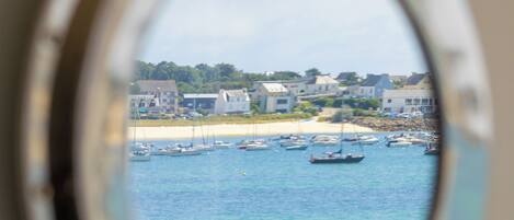 A view through a circular window of a coastal area with boats, buildings, and a sandy beach.