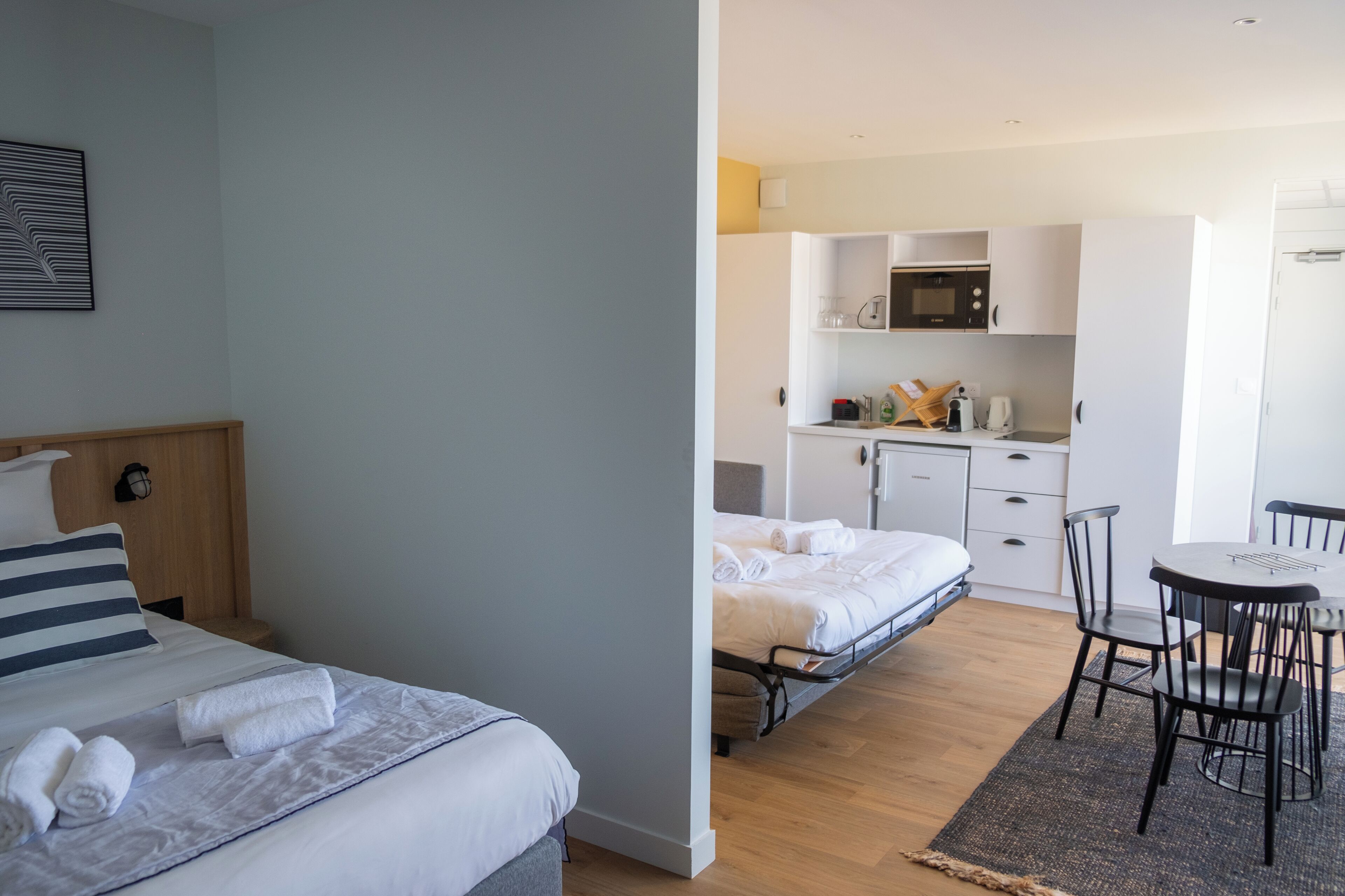 A room with two beds, a dining table with chairs, a kitchen area with a microwave, and a small cabinet.