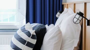 Una cama bien hecha con almohadas blancas y a rayas, un taburete tejido pequeño y una ventana con cortinas.