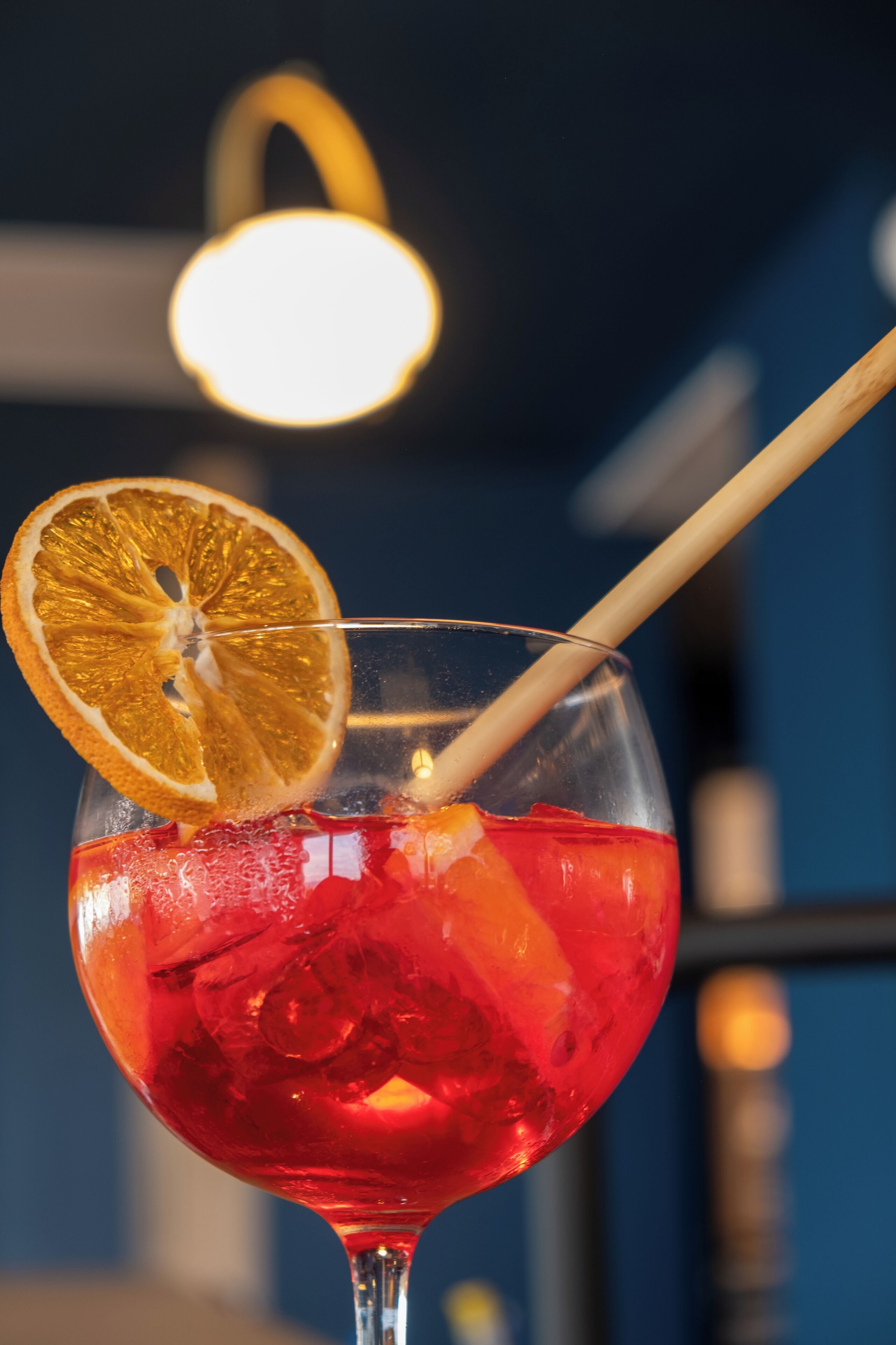 Un vaso de cóctel con una rodaja de naranja y una pajita, lleno de una bebida roja y cubitos de hielo.