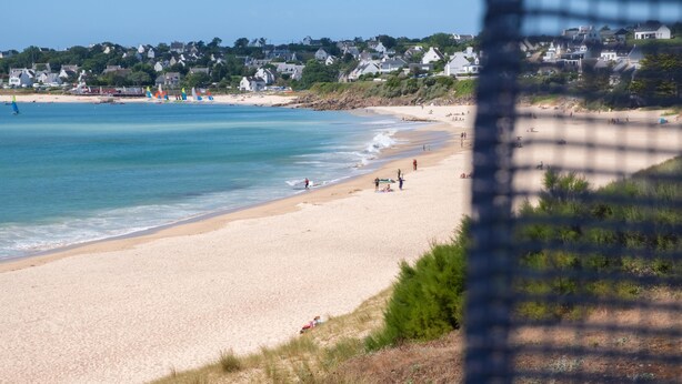 Une plage avec une eau d’un bleu limpide, un rivage sablonneux et des maisons en arrière-plan.