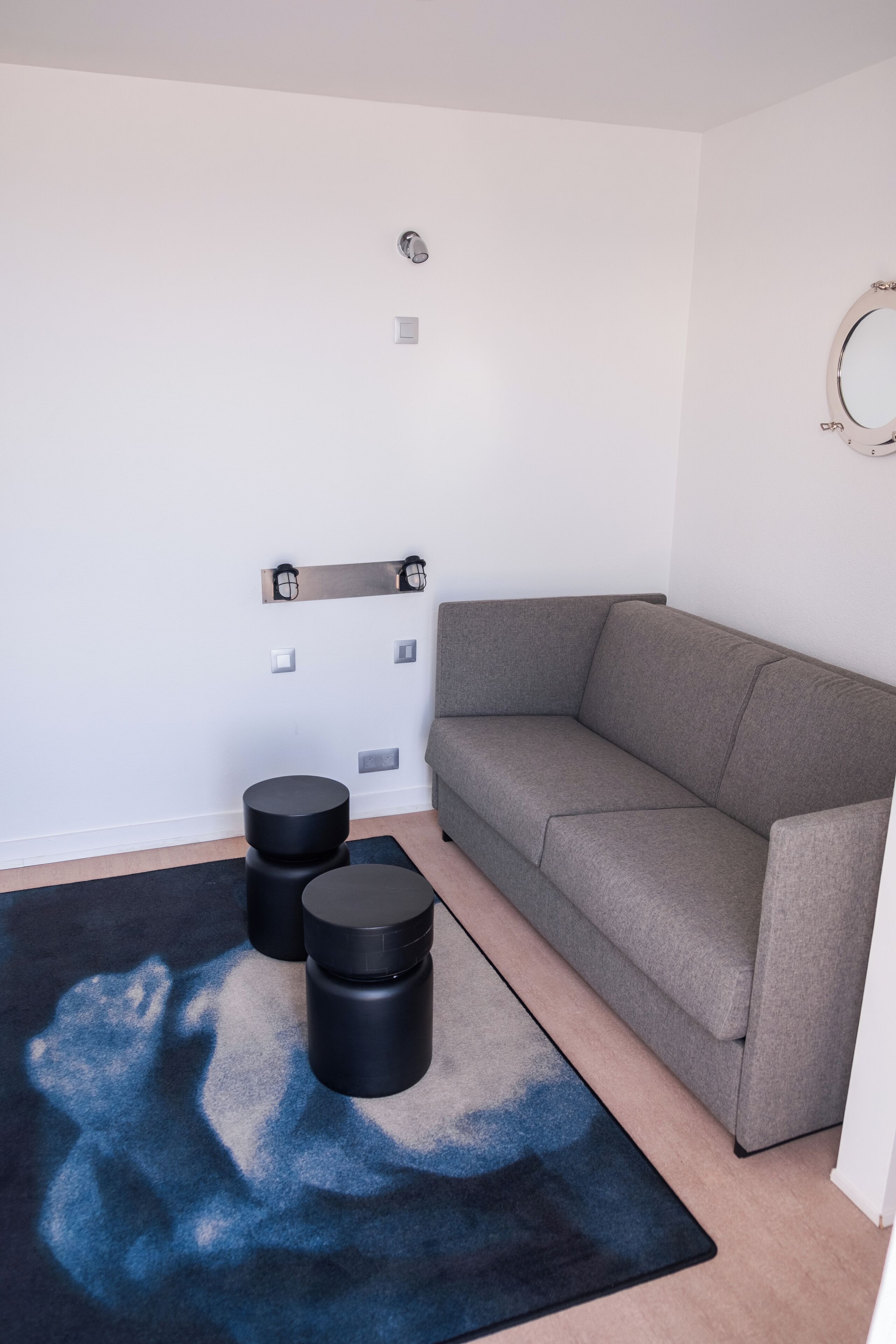 Un salon moderne avec un canapé gris, un miroir rond et deux poufs noirs posés sur un tapis bleu et noir.